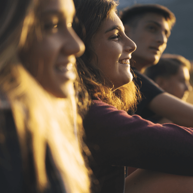 Four teenagers sitting down in a field at sunset, talking and laughing.